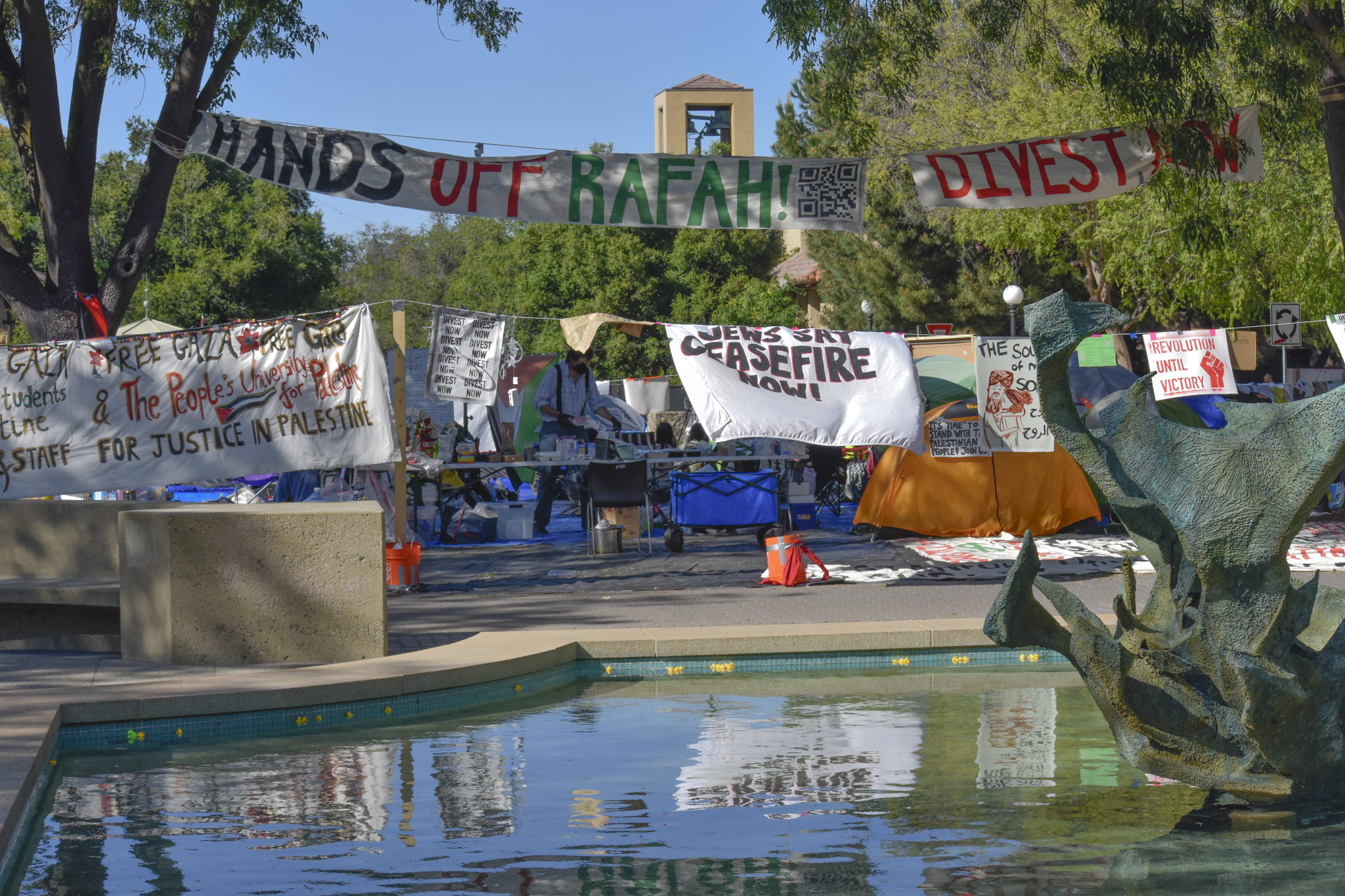 The student encampment at White Plaza. Standing in front of the White Memorial fountain is an encampment surrounded by tents and an outer layer of banners. One sign states “Revolution Until Victory.”